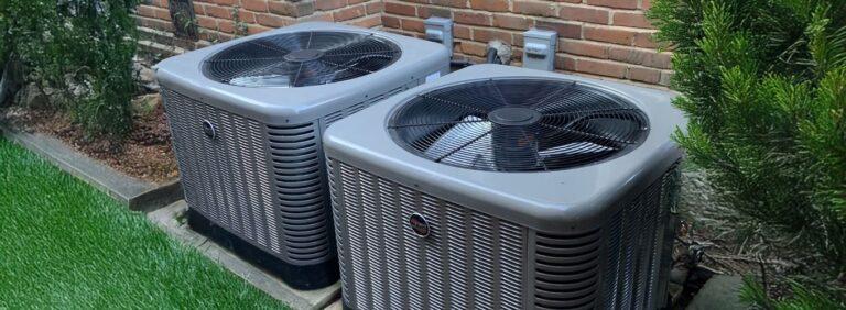 Two residential outdoor AC condenser units positioned beside a brick wall, surrounded by landscaping