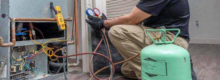 Licensed HVAC technician servicing indoor AC unit with refrigerant gauges and recovery tank during refrigerant recharge