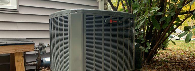 Outdoor AC condenser unit beside a home with nearby plants and siding
