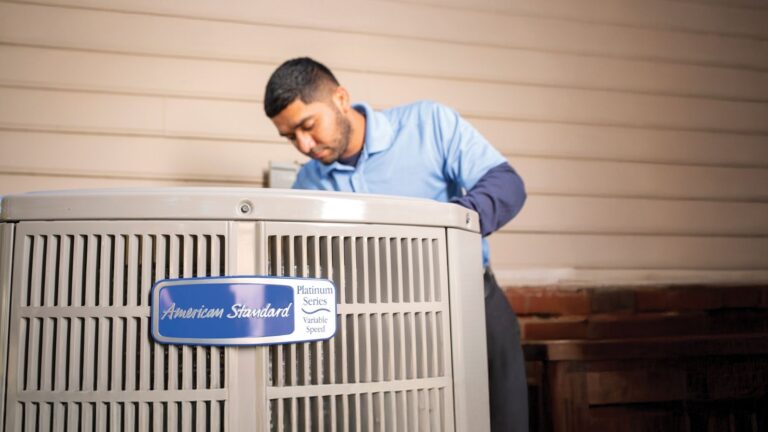 HVAC technician servicing an American Standard air conditioner unit outside a home.