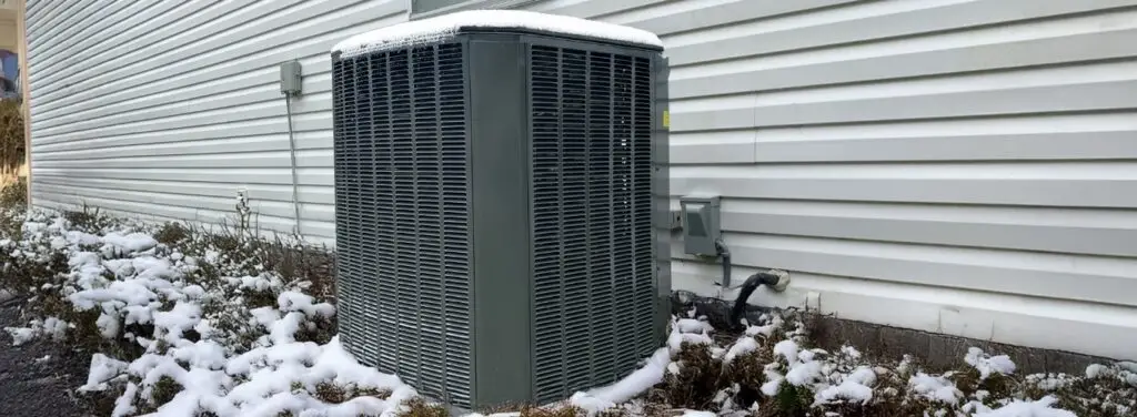 Outdoor HVAC unit and surrounding bushes lightly covered in snow against the white siding of a house during winter.
