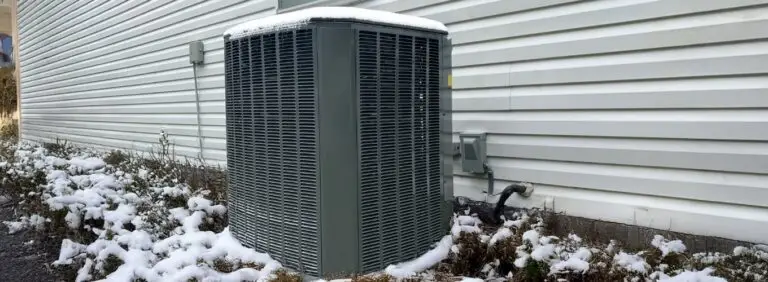 Outdoor HVAC unit and surrounding bushes lightly covered in snow against the white siding of a house during winter.