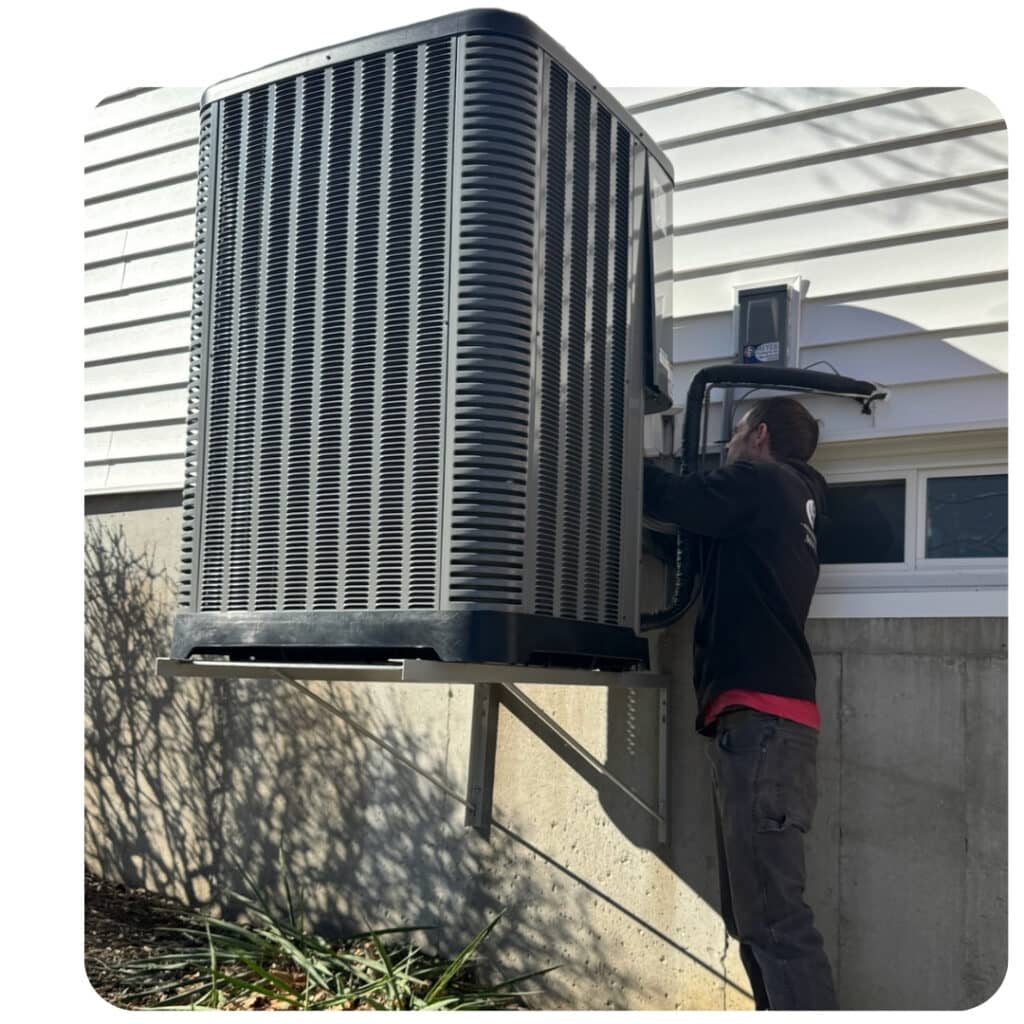 A service technician working on an elevated air conditioner condenser unit secured to an exterior wall.