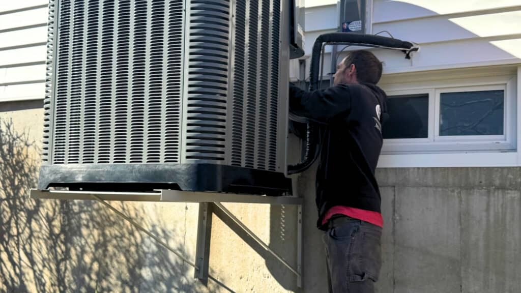 Meyer Heating & Air technician performing seasonal maintenance on an elevated, wall-bracketed outdoor heat pump unit.