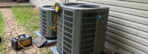 Two gray American Standard outdoor HVAC condenser units being serviced with digital diagnostic tools in a backyard.
