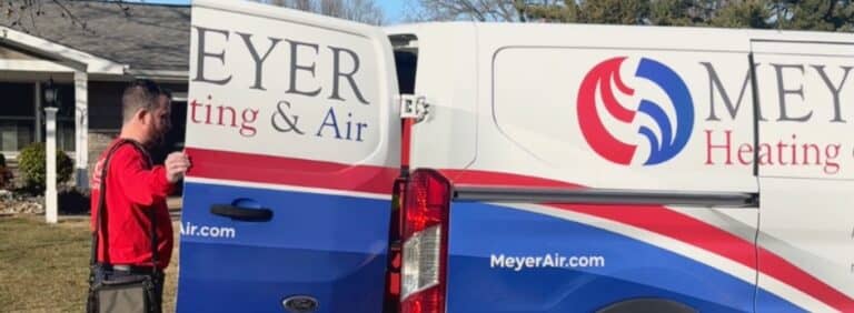 Local HVAC technician in a red uniform shirt opening the back doors of a white Meyer Heating & Air service van parked in a driveway.