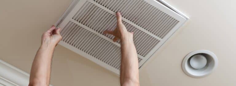 Homeowner replacing a dirty return air vent filter in the ceiling to prevent furnace airflow issues and cold air blowing.